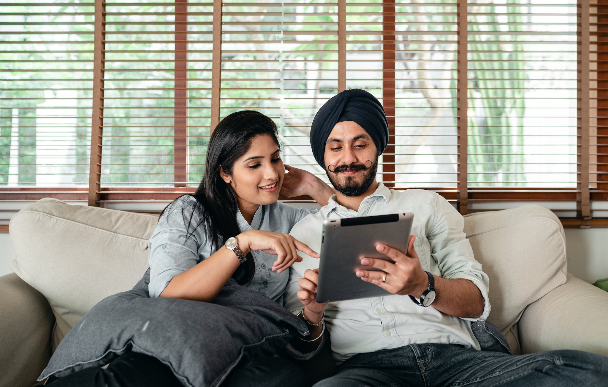 Content Indian woman and man in turban resting on cozy couch in living room and sharing tablet